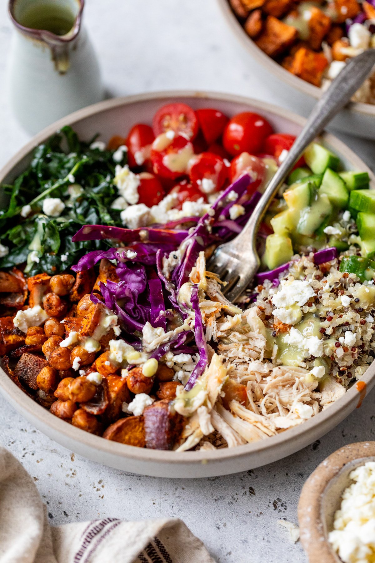 Eine lebendige grüne Göttin -Schüssel mit gesunden Zutaten, einschließlich zerkleinertem Hühnchen, gerösteten Kichererbsen, Quinoa, Gurken, Kirschtomaten, Grünkohl, Süßkartoffeln, rotem Kohl und zerbröckeltem Feta -Käse. Eine Gabel ruht in der Schüssel.