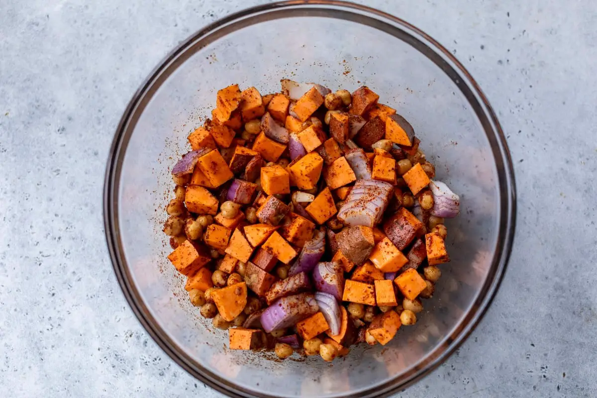 Auf einer hellgrauen Arbeitsplatte sitzt eine Glasschüssel mit der herzhaften Güte gewürzter Süßkartoffeln, Kichererbsen und roten Zwiebeln, die perfekt gewürzt sind. Dieses entzückende Gericht fängt die Essenz der grünen Göttin -Schalen in jedem Bissen ein.