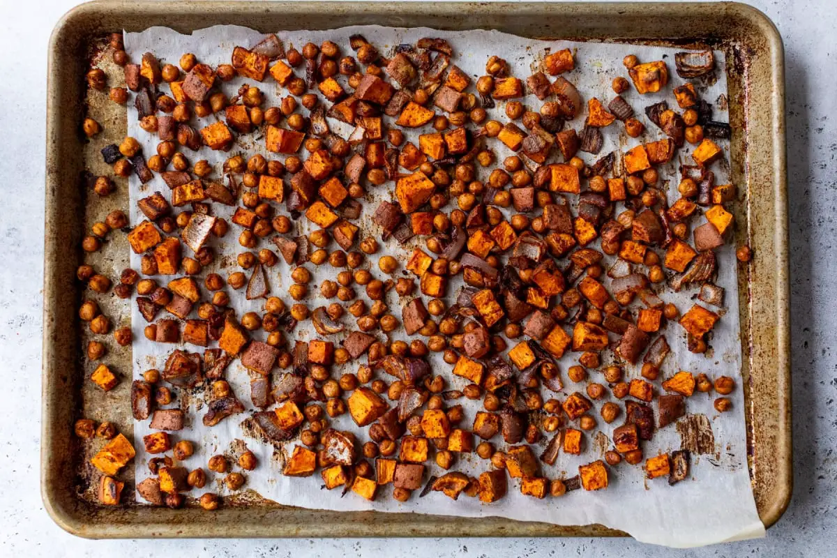 Gebratenes Gemüse und Kichererbsen auf einem mit Pergament ausgekleideten Backblech, perfekt für grüne Göttinschalen. Die Mischung umfasst gewürfelte Süßkartoffeln, rote Zwiebeln und Kichererbsen, die alle gewürzt und gebräunt sind. Das Blatt hat einen rustikalen Look mit leichtem Verkohlung am Gemüse.