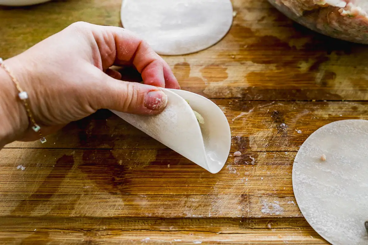 Eine Hand hält eine teilweise gefaltete kreisförmige Knödelhülle auf einer Holzoberfläche. In der Nähe befinden sich mehrere nicht gefüllte Wrapper, und eine Schüssel mit Füllung ist im Hintergrund teilweise sichtbar.