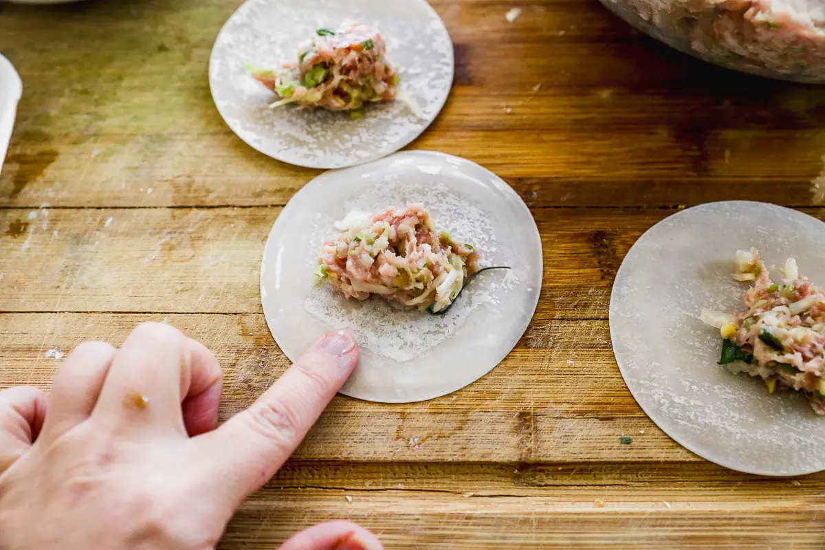 Nahaufnahme einer Hand, die Knödel auf einer Holzoberfläche montiert. Kreisförmige Knödelverpackungen sind mit einer Mischung aus gehacktem Fleisch und Gemüse gefüllt. Eine Verpackung wird zum Versiegeln mit einem Finger vor seiner Kante vorbereitet.