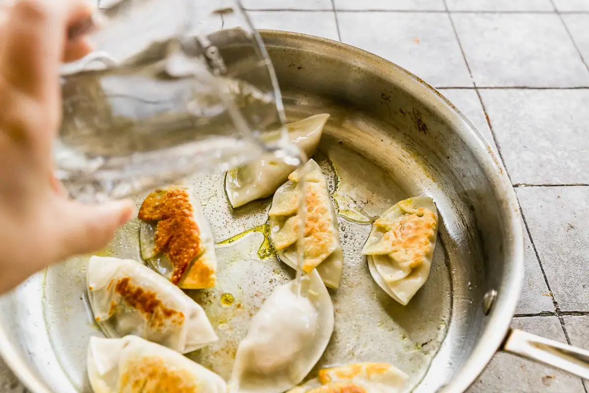 Eine Hand gießt Wasser in eine Pfanne mit brutzelnden Schweinefleischknödeln. Die Knödel sind auf einer Seite goldbraun und die Pfanne ist auf einer gefliesten Oberfläche eingestellt.