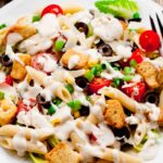 A plate of Caesar pasta salad with penne, cherry tomatoes, black olives, croutons, green onions, and lettuce, topped with a creamy dressing.