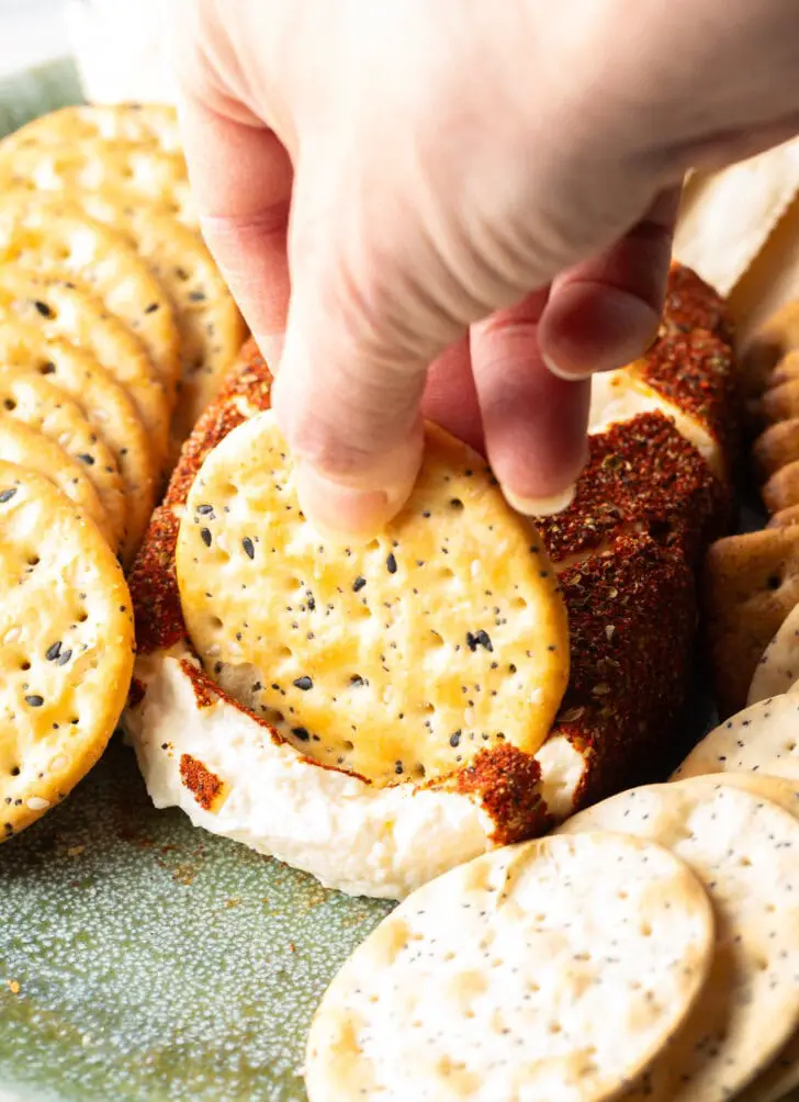 Hand mit einem Cracker schöpft eine Portion geräucherten Frischkäse aus dem Block, umgeben von Crackern.