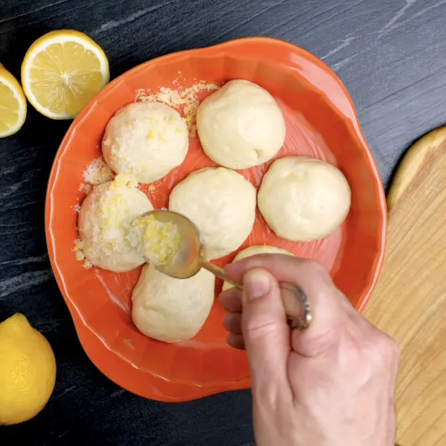 Eine Hand streut eine Mischung aus Zitronenschale und Zucker über sieben runde Teigbällchen in einer Auflaufform. 