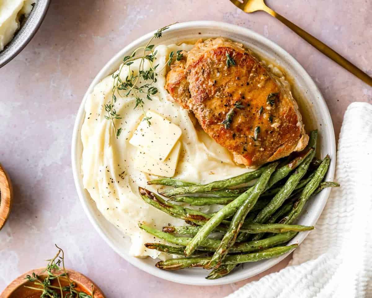 Draufsicht auf ein im Crockpot zubereitetes Schweinekotelett auf einem weißen Teller mit grünen Bohnen und Kartoffelpüree.