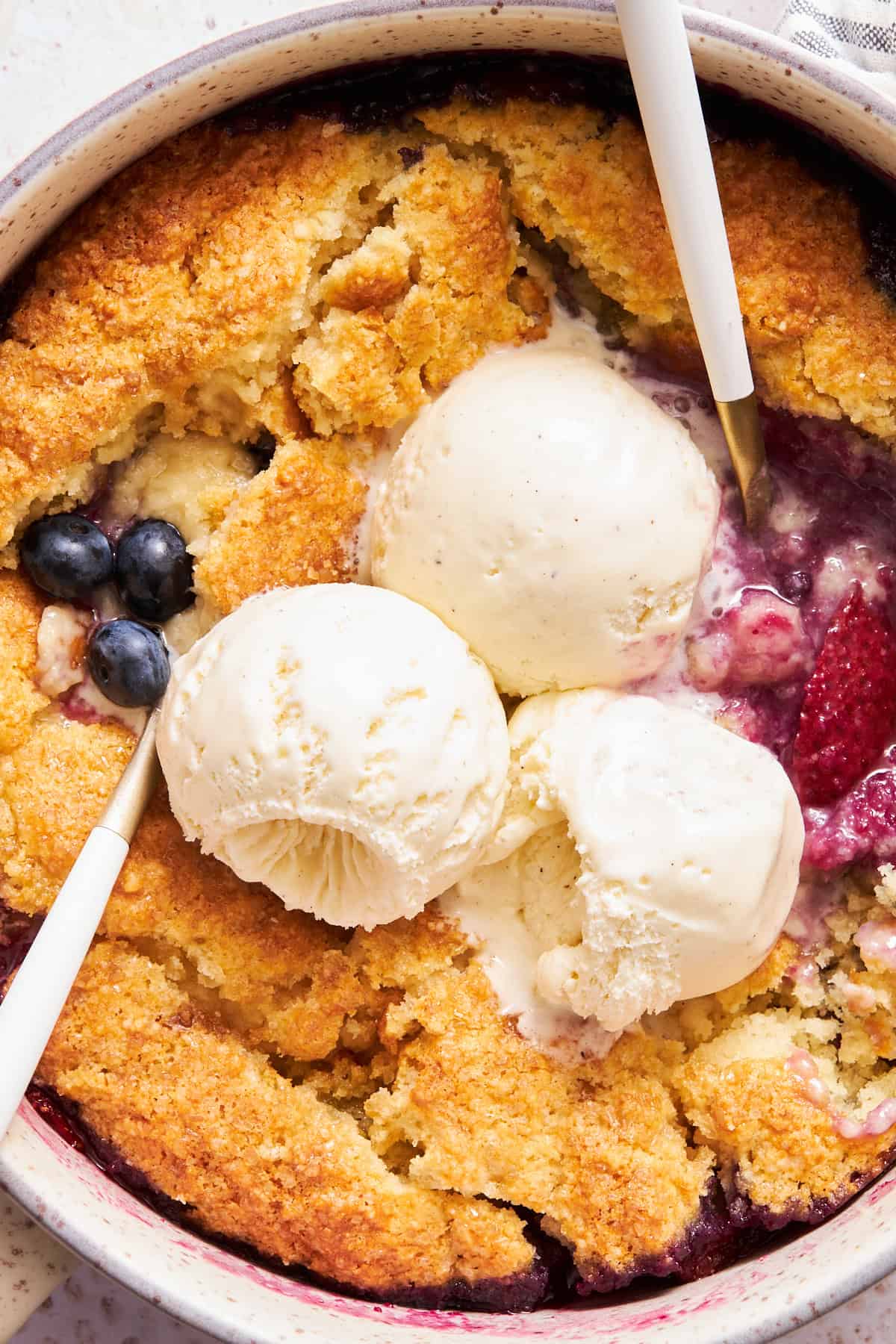 Nahaufnahme von zwei Löffeln in einem Cobbler aus gemischten Beeren, garniert mit drei Kugeln Eis. 