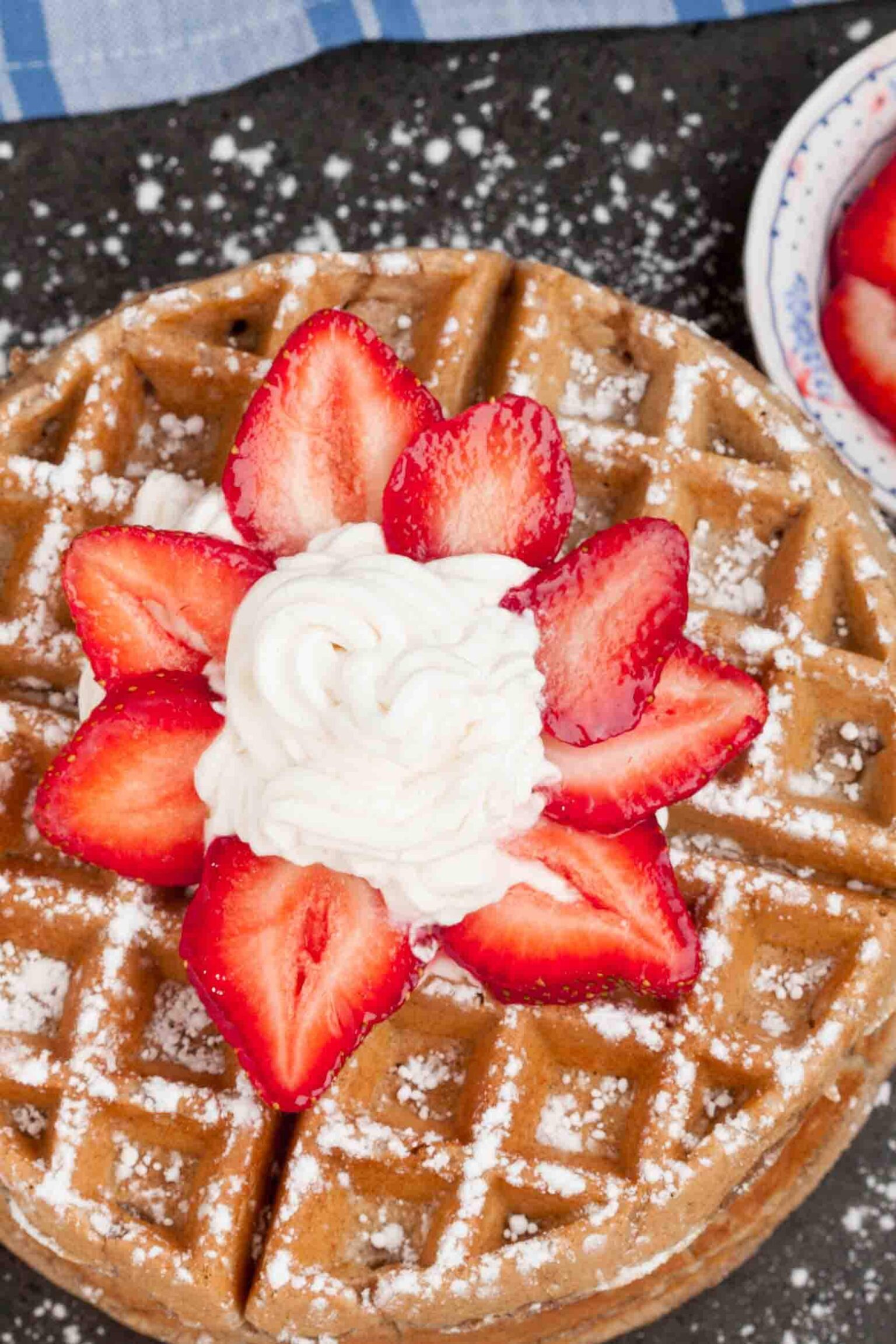 A close-up of a waffle topped with powdered sugar, whipped cream, and sliced strawberries arranged in a flower shape.