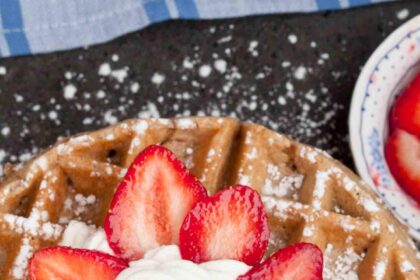 A close-up of a waffle topped with powdered sugar, whipped cream, and sliced strawberries arranged in a flower shape.