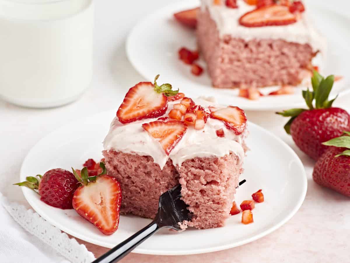 Seitenansicht eines Stücks Erdbeerblechkuchen auf einem kleinen weißen Teller, in dessen Mitte eine Gabel eingeschnitten ist.