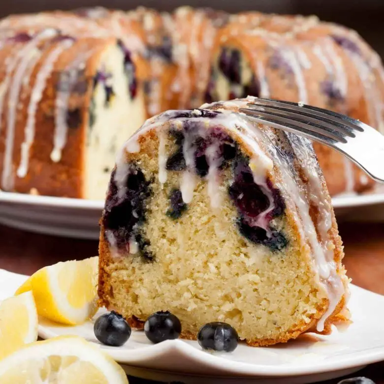 Ein Stück Blaubeer-Zitronen-Gugelhupf mit Zuckerguss auf einem Teller, garniert mit Zitronenscheiben und Blaubeeren. 