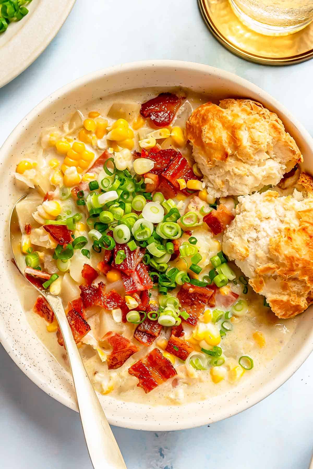 two biscuits are in the bowl of corn chowder with a metal spoon and topped with green onions and chopped bacon