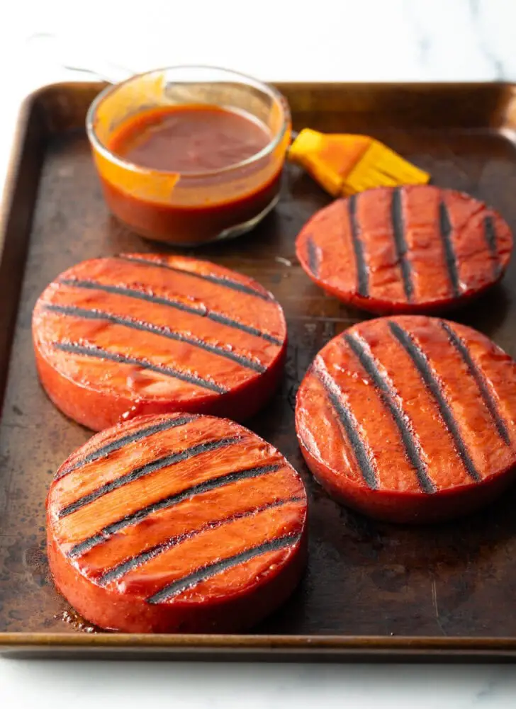 4 Scheiben Bologna mit Grillstreifen. Im Hintergrund in einer Glasschale mit Barbecuesauce und einem gelben Backpinsel.