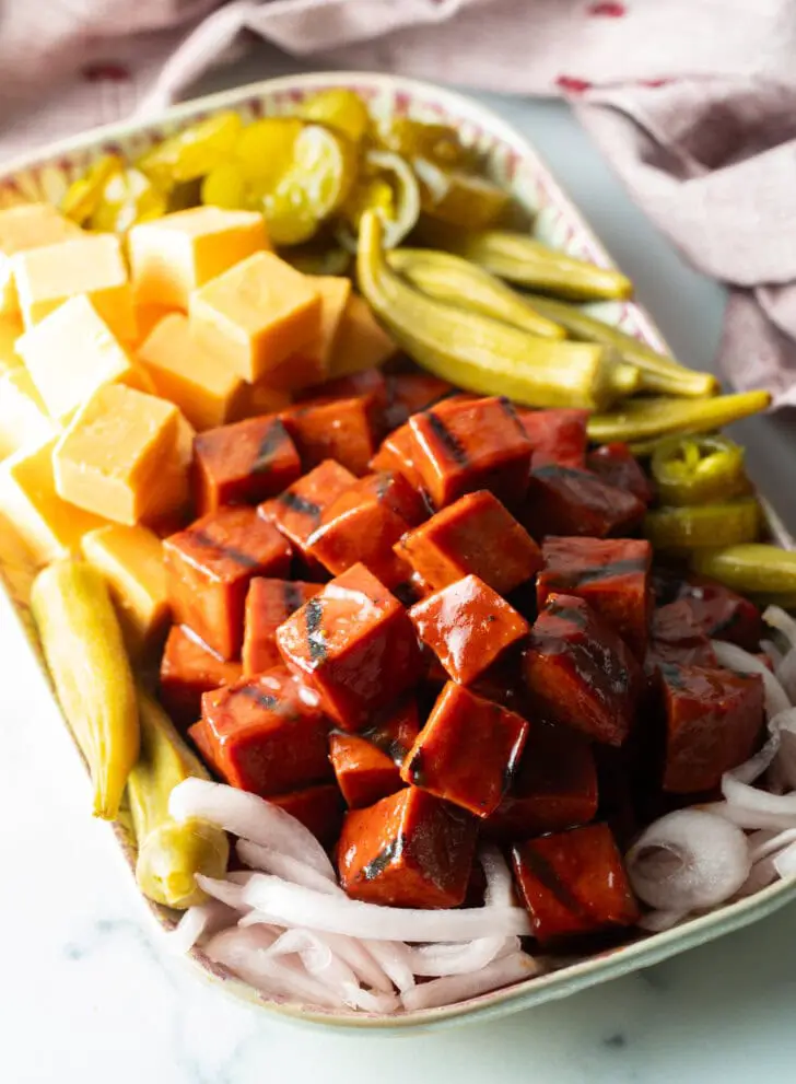 Servierteller mit einem Stapel geräucherter BBQ-Würfel aus Bologna, Käsequadraten, eingelegter Okraschoten, roten Zwiebelscheiben und Gewürzgurken.