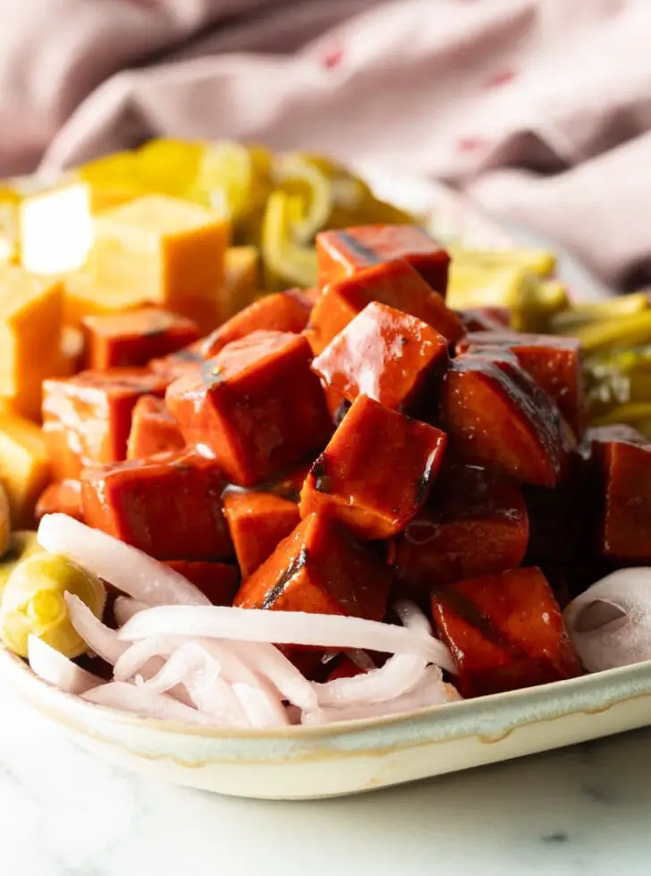 Servierteller mit einem Stapel geräucherter BBQ-Würfel aus Bologna, Käsequadraten, eingelegter Okraschoten, roten Zwiebelscheiben und Gewürzgurken.