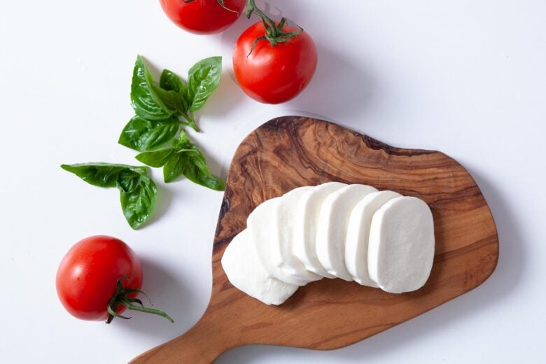 in Scheiben geschnittener frischer Mozzarella auf einem Holzschneidebrett mit Tomaten und Basilikum an der Seite