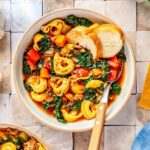 a bowl with a metal spoon has sausage tortellini soup and sliced bread on the inside