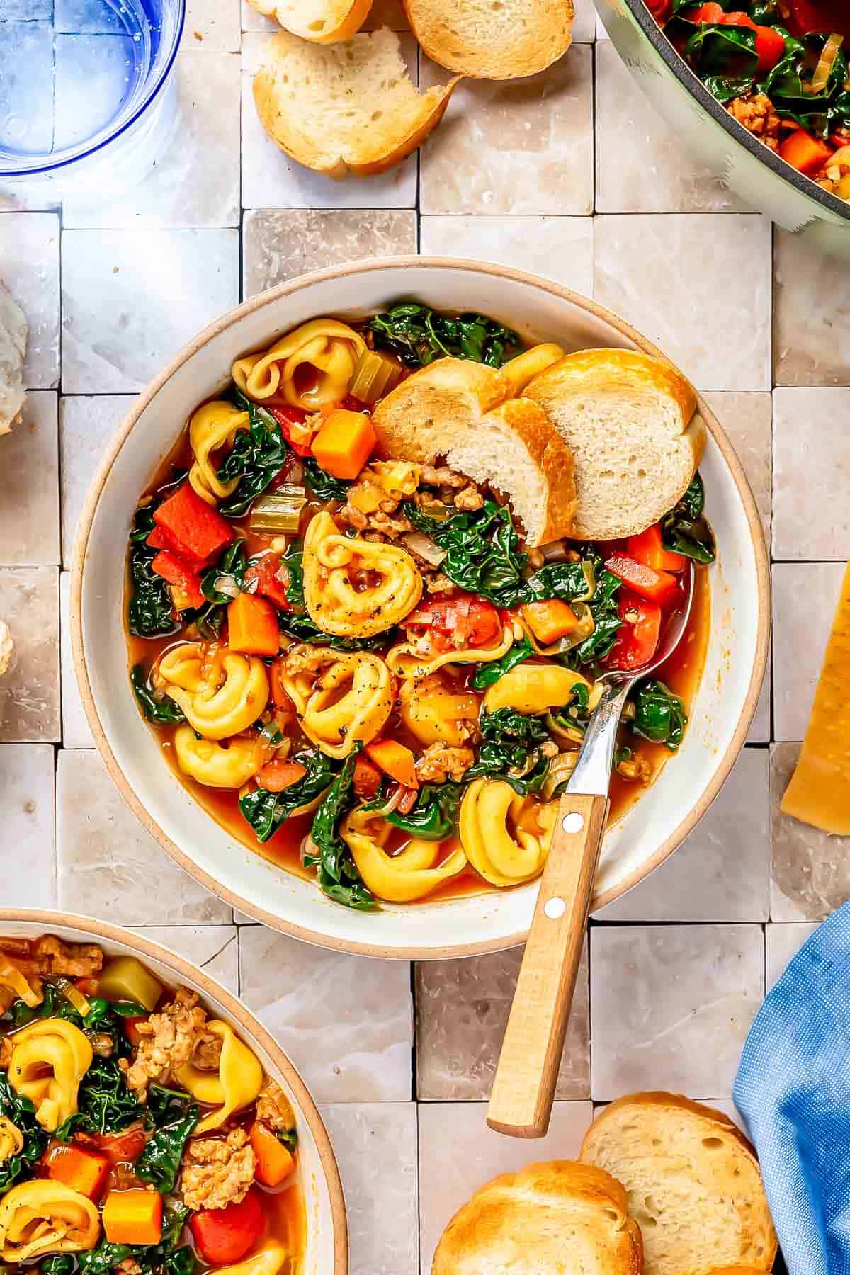 a bowl with a metal spoon has sausage tortellini soup and sliced bread on the inside