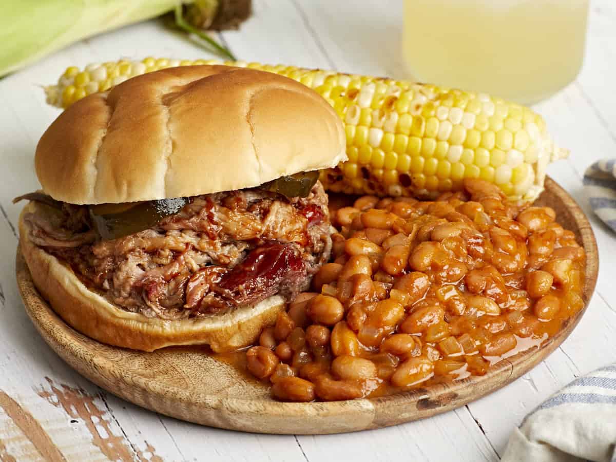 eine Portion gebackene Bohnen auf einem Teller mit einem Pulled-Pork-Sandwich und Maiskolben.