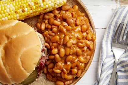 overhead view of a serving of baked beans on a plate with a sandwich and corn on the cob.