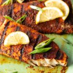 Close view of a fillet of grilled mahi mahi topped with a slice of lemon and green onions. A bite has been removed from the fish to show the white flaky inside of the meat.