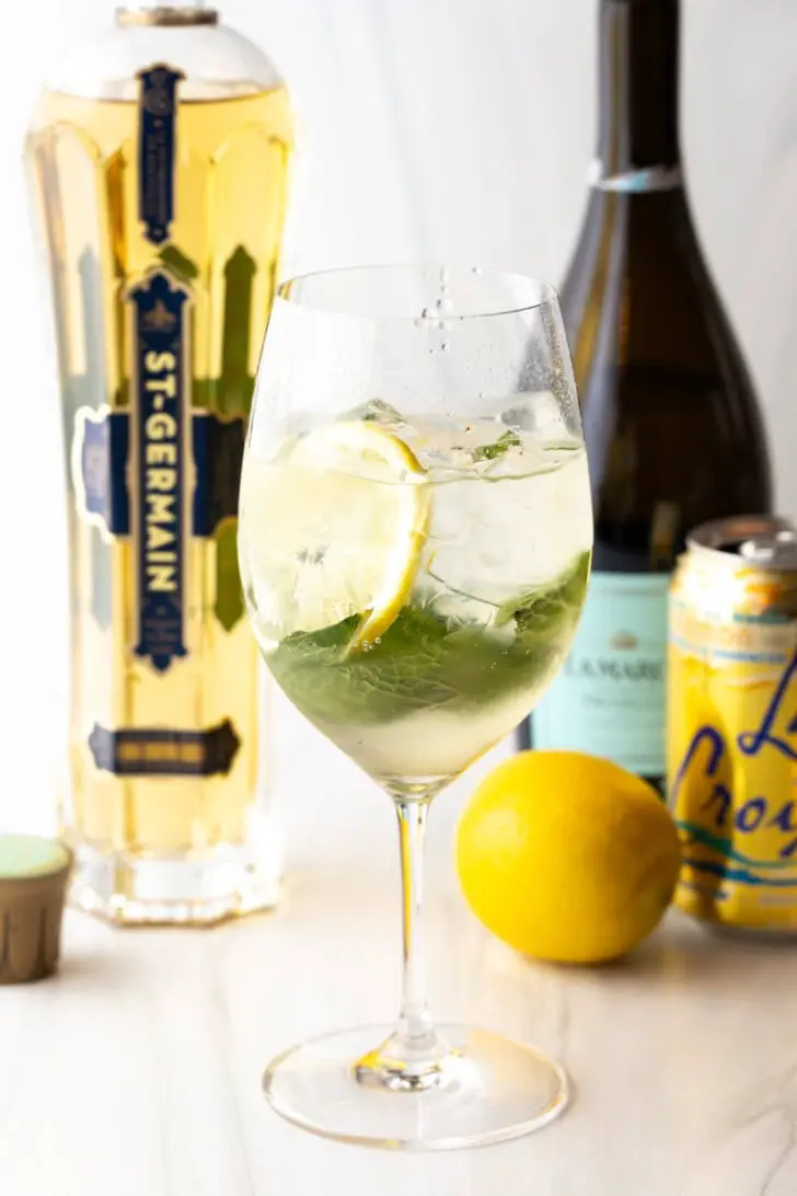 Weinglas mit Alkohol, Minzblättern und Zitronenscheiben. Im Hintergrund des Hugo Spritz sind eine ganze Zitrone und Flaschen Prosecco, St. Germain-Likör und eine Dose La Croix-Limonade zu sehen.