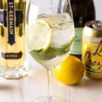 Wine glass with alcohol, mint leaves, and lemon slices. In the background of the hugo spritz are a whole lemon and bottles of prosecco, st. germain liquor, and a can of la croix soda.