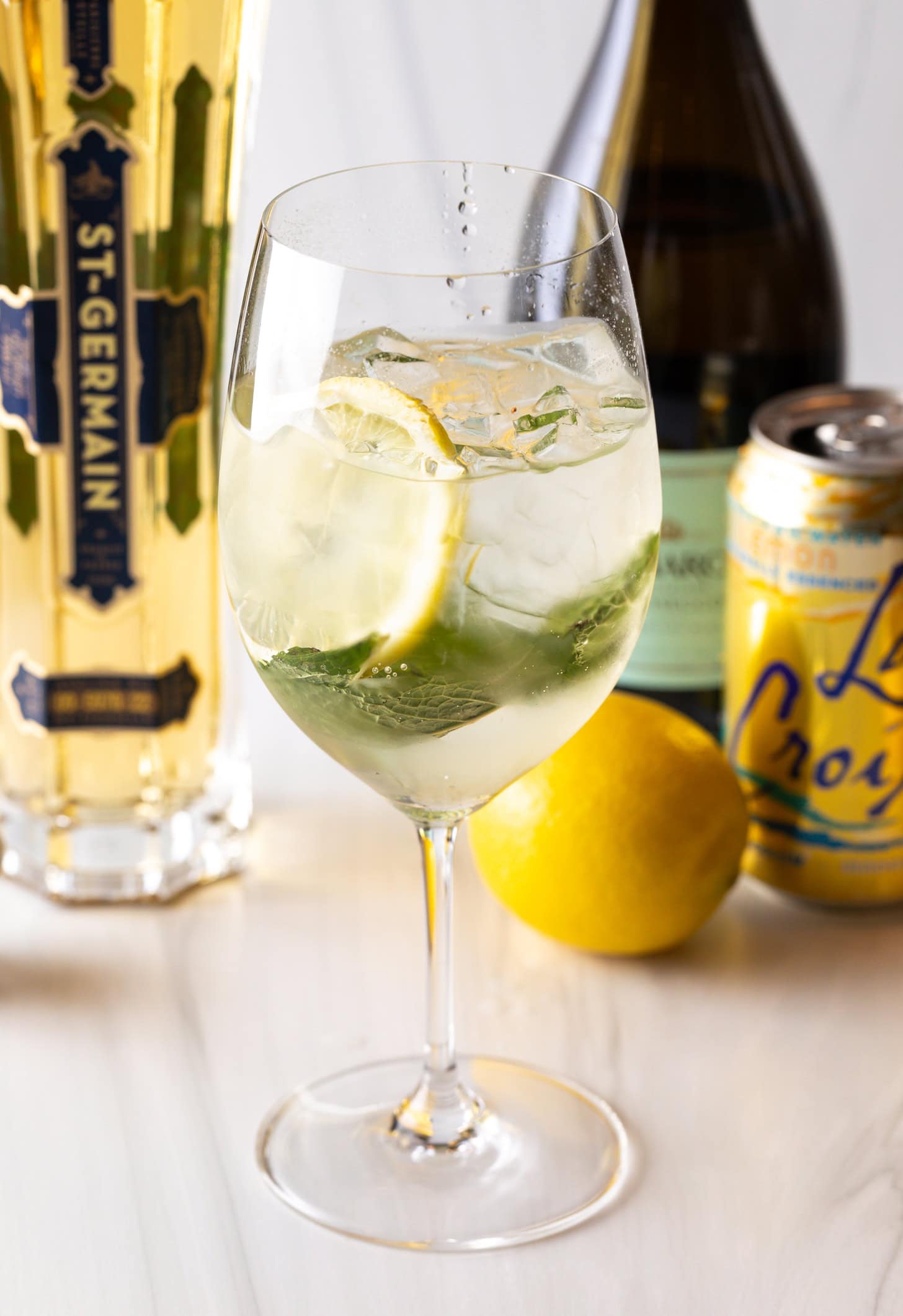 Wine glass with alcohol, mint leaves, and lemon slices. In the background of the hugo spritz are a whole lemon and bottles of prosecco, st. germain liquor, and a can of la croix soda.