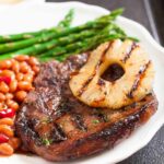 A plate with grilled pork chops with marinade topped with a grilled pineapple slice, accompanied by baked beans and asparagus.