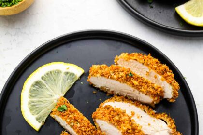 a sliced panko breaded chicken breast on a black plate with lemon wedges.