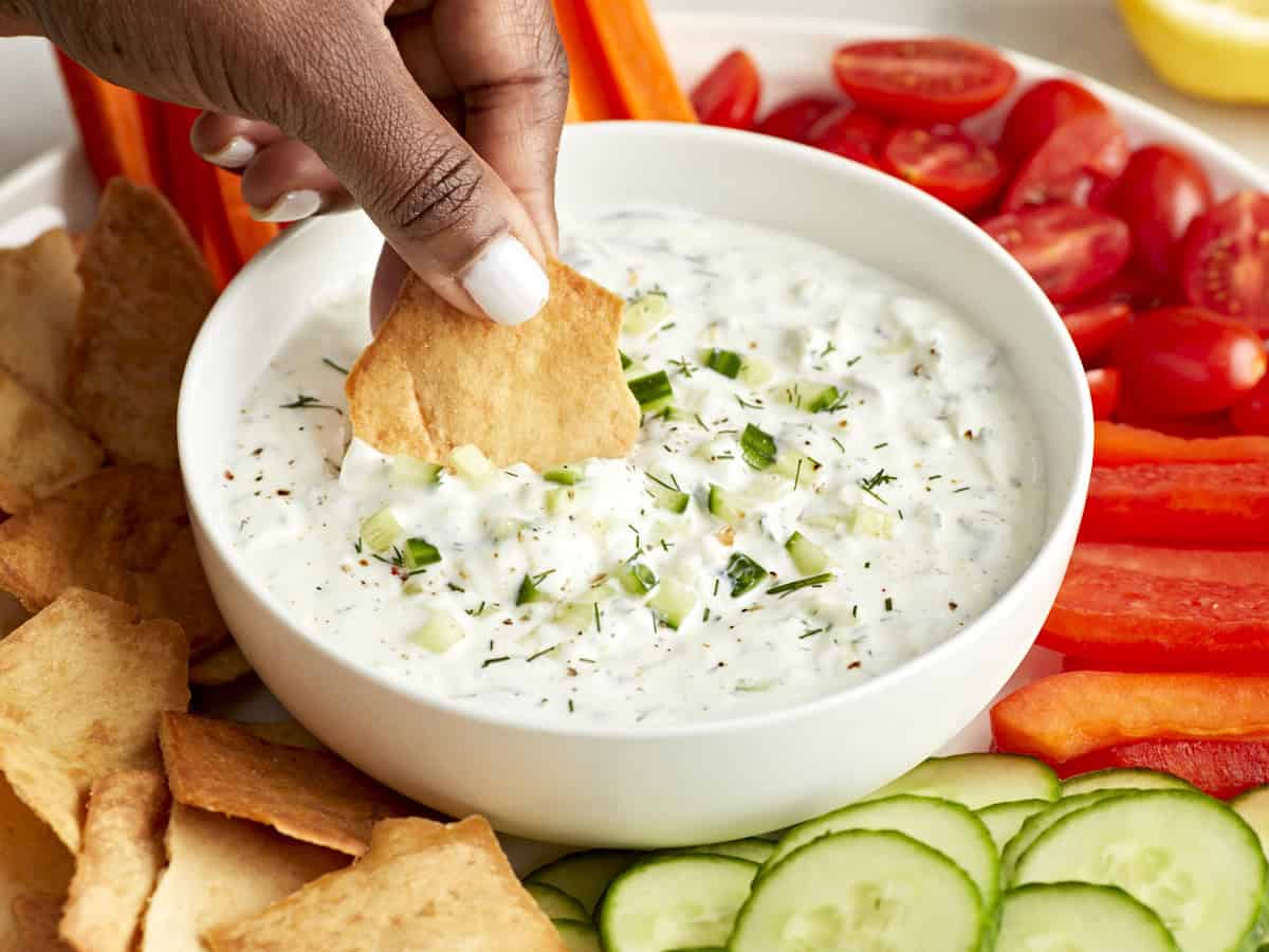 Seitenansicht einer Tzatziki-Sauce in einer weißen Servierplatte mit einem in die Sauce getauchten Pita-Chip.