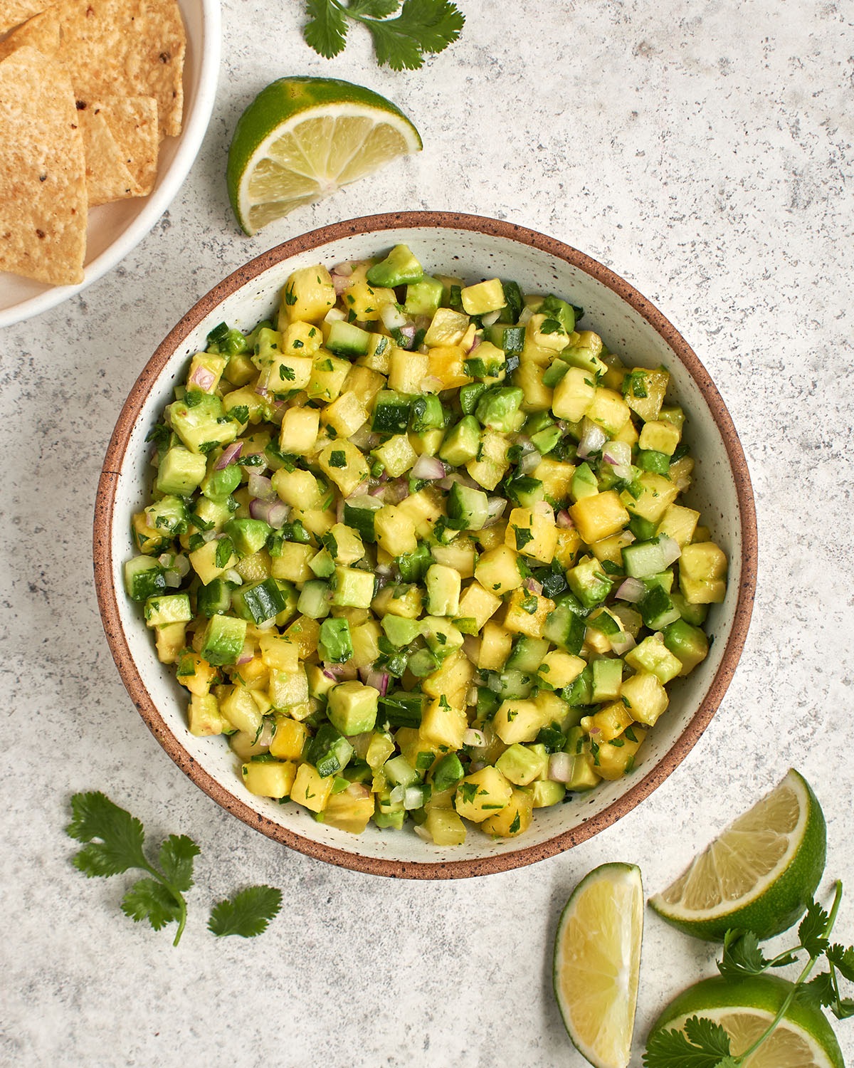 Ananas-Avocado-Salsa in einer Schüssel, umgeben von Limettenspalten und einer weißen Schüssel mit Tortillachips