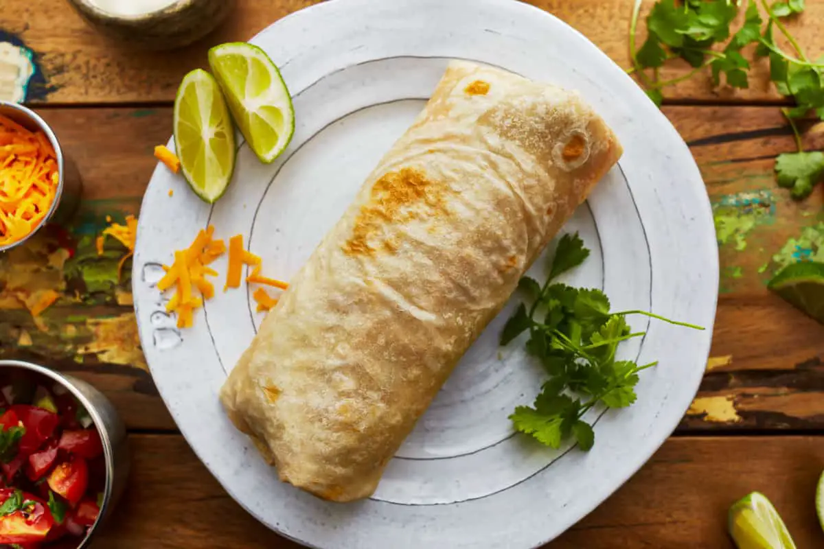 Ein Burrito auf einem weißen Teller mit Limetten und Koriander.