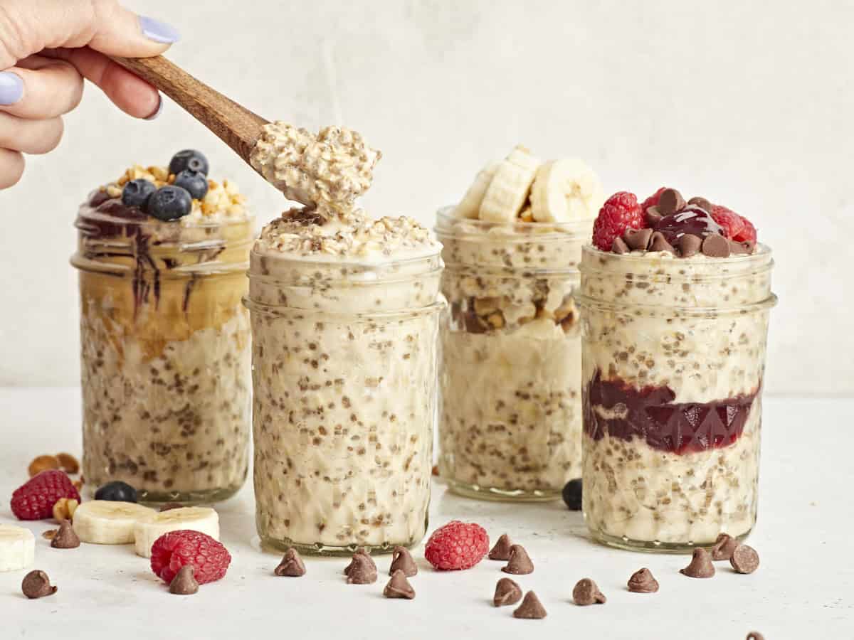 Seitenansicht von 4 Gläsern Overnight Oats mit einem Holzlöffel, aus einem der Gläser wird etwas entnommen