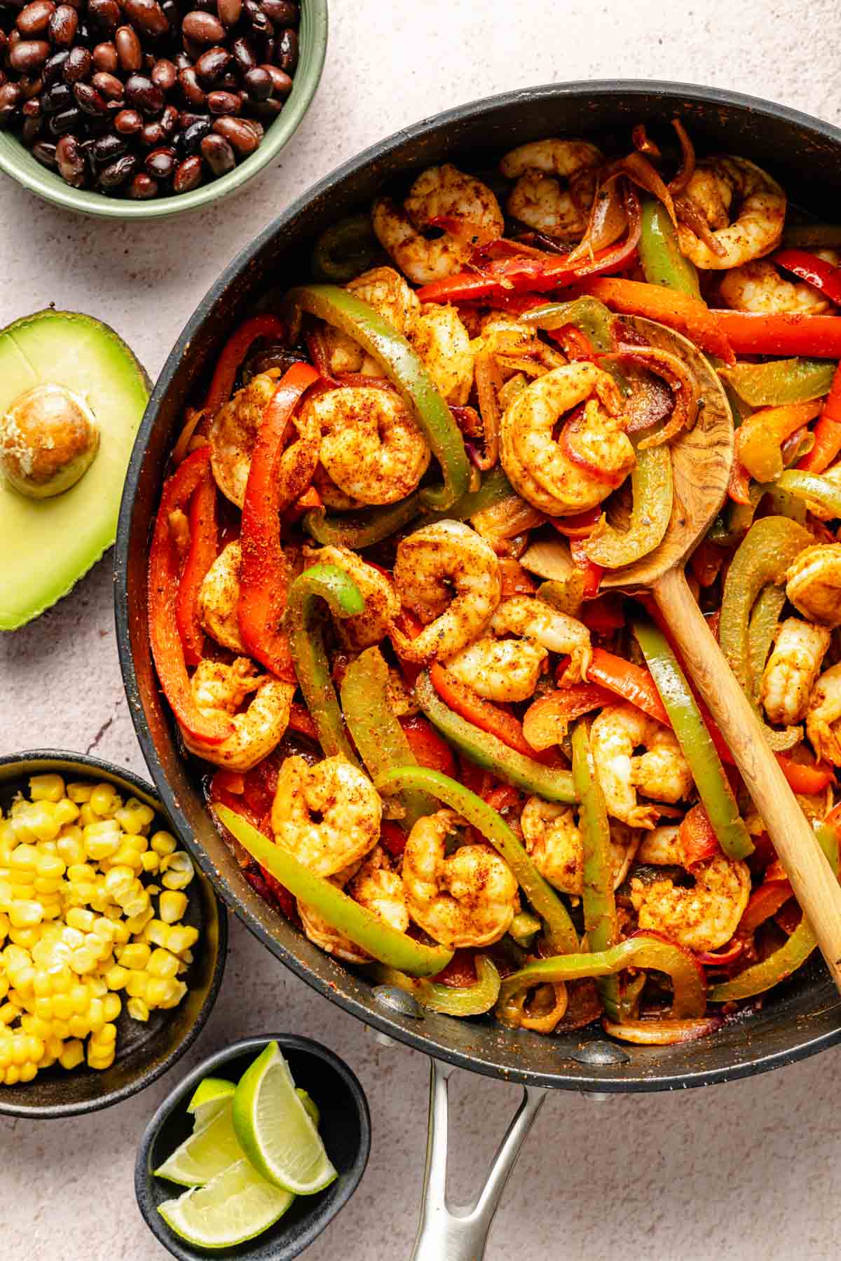 Eine Pfanne ist mit Garnelen, geschnittenen Paprika und Zwiebeln gefüllt, mit einem Holzlöffel neben frischen Zutaten wie Limettenschnitzen, Mais, schwarzen Bohnen und Avocado