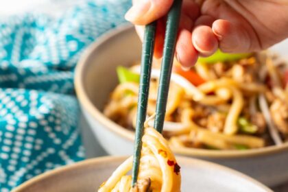 Hand with chopsticks grabbing some noodles from the bowl of stir fried vegetables and chicken.