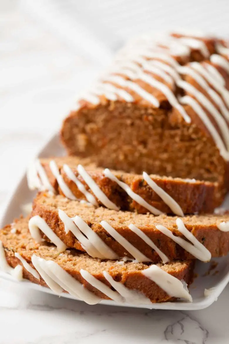 Ein Laib teilweise aufgeschnittenes Zucchini-Karottenbrot mit Frischkäse-Zuckerguss auf einer weißen Oberfläche.