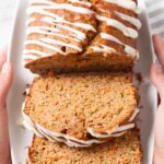 A plate with sliced zucchini carrot bread drizzled with cream cheese frosting, being held by two hands.