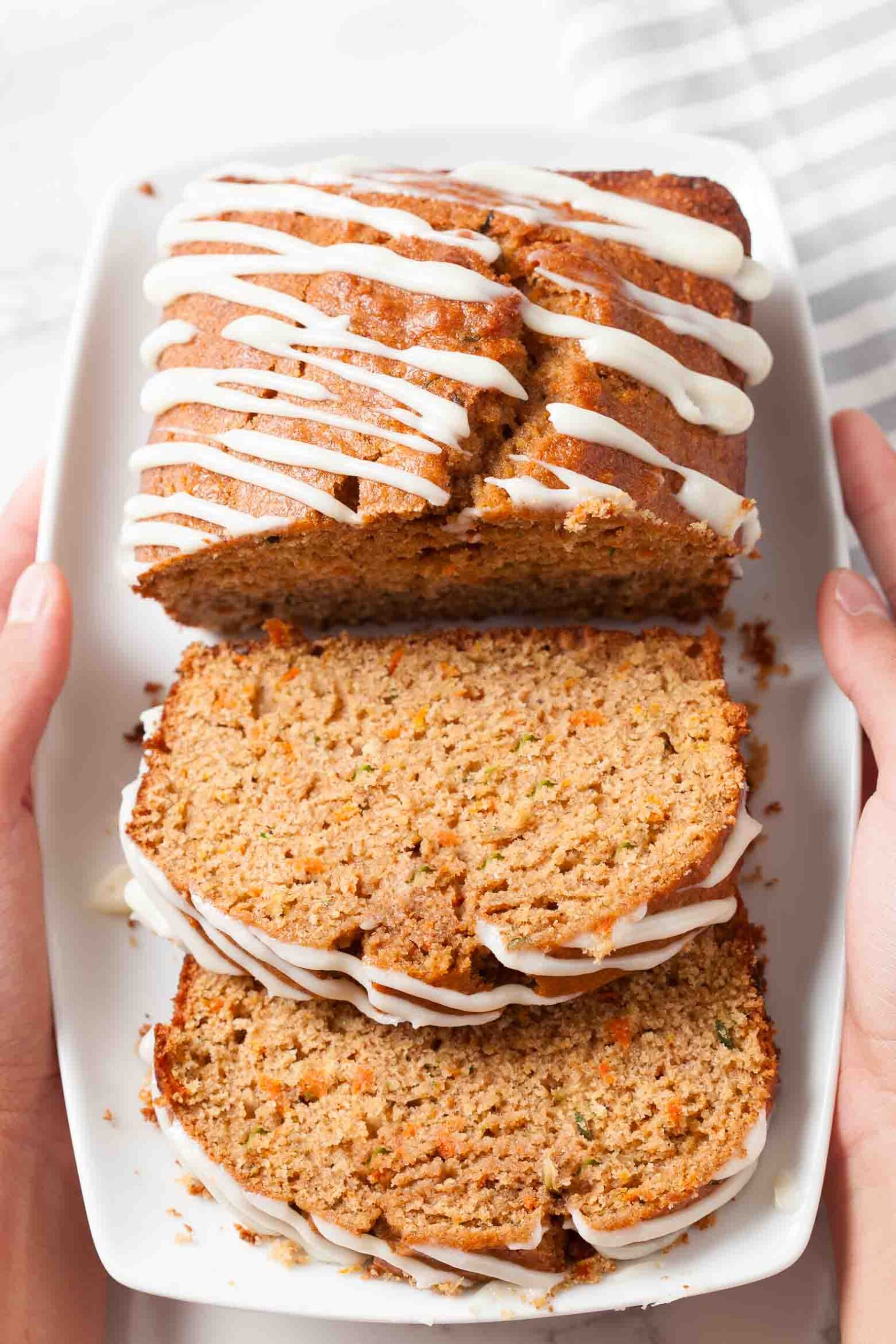 A plate with sliced zucchini carrot bread drizzled with cream cheese frosting, being held by two hands.