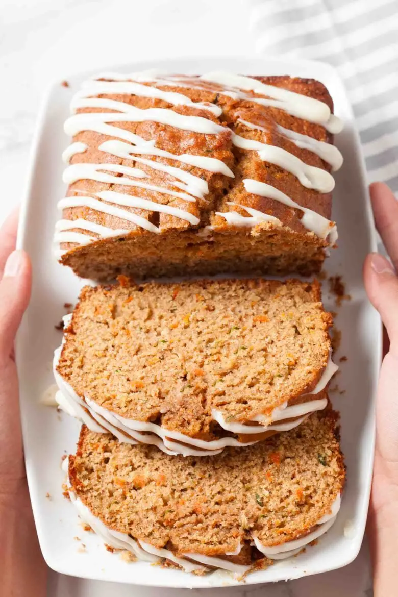 Ein Teller mit geschnittenem Zucchini-Karottenbrot, beträufelt mit Frischkäse-Zuckerguss, wird von beiden Händen gehalten.