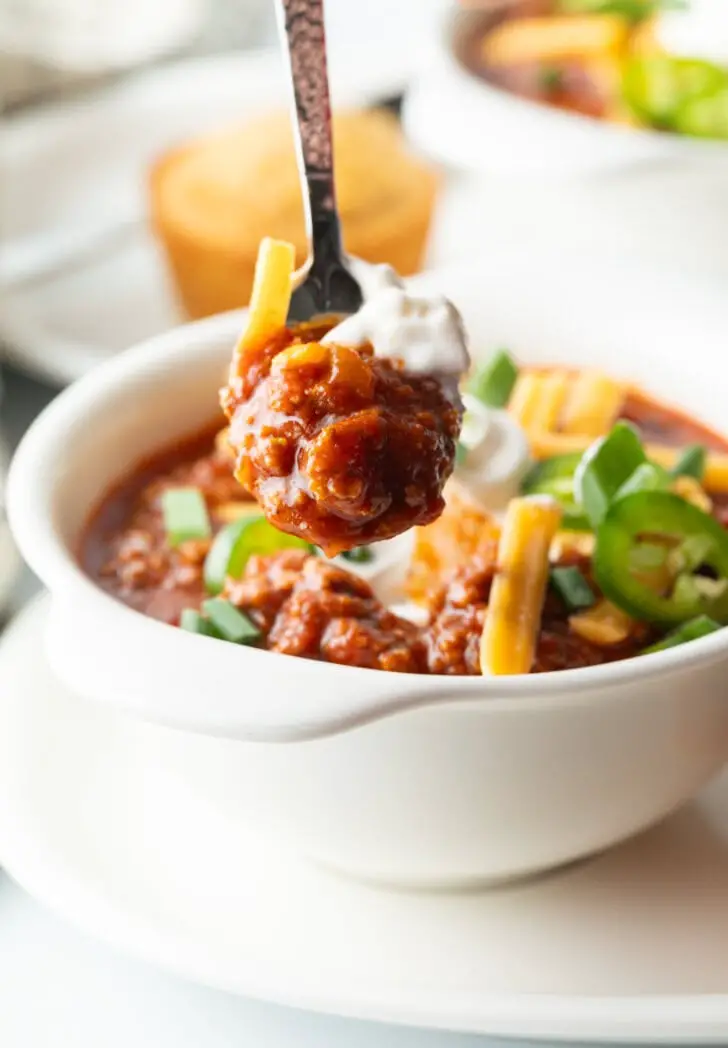 Weiße Schüssel Chili ohne Bohnen, garniert mit geriebenem Käse, Jalapeno-Scheiben, Sauerrahm und gehackten Frühlingszwiebeln. Ein Löffel zeigt einen Löffel Chili con Carne in die Kamera.