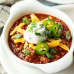 Top down view of a white bowl of beanless chili topped with shredded cheese, jalapeno slices, sour cream and chopped green onions.