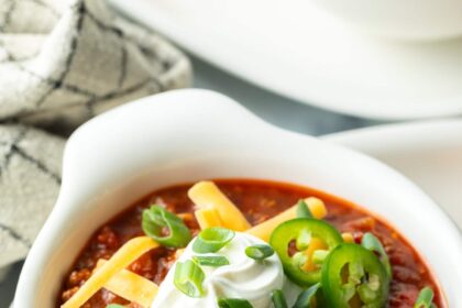 Top down view of a white bowl of beanless chili topped with shredded cheese, jalapeno slices, sour cream and chopped green onions.