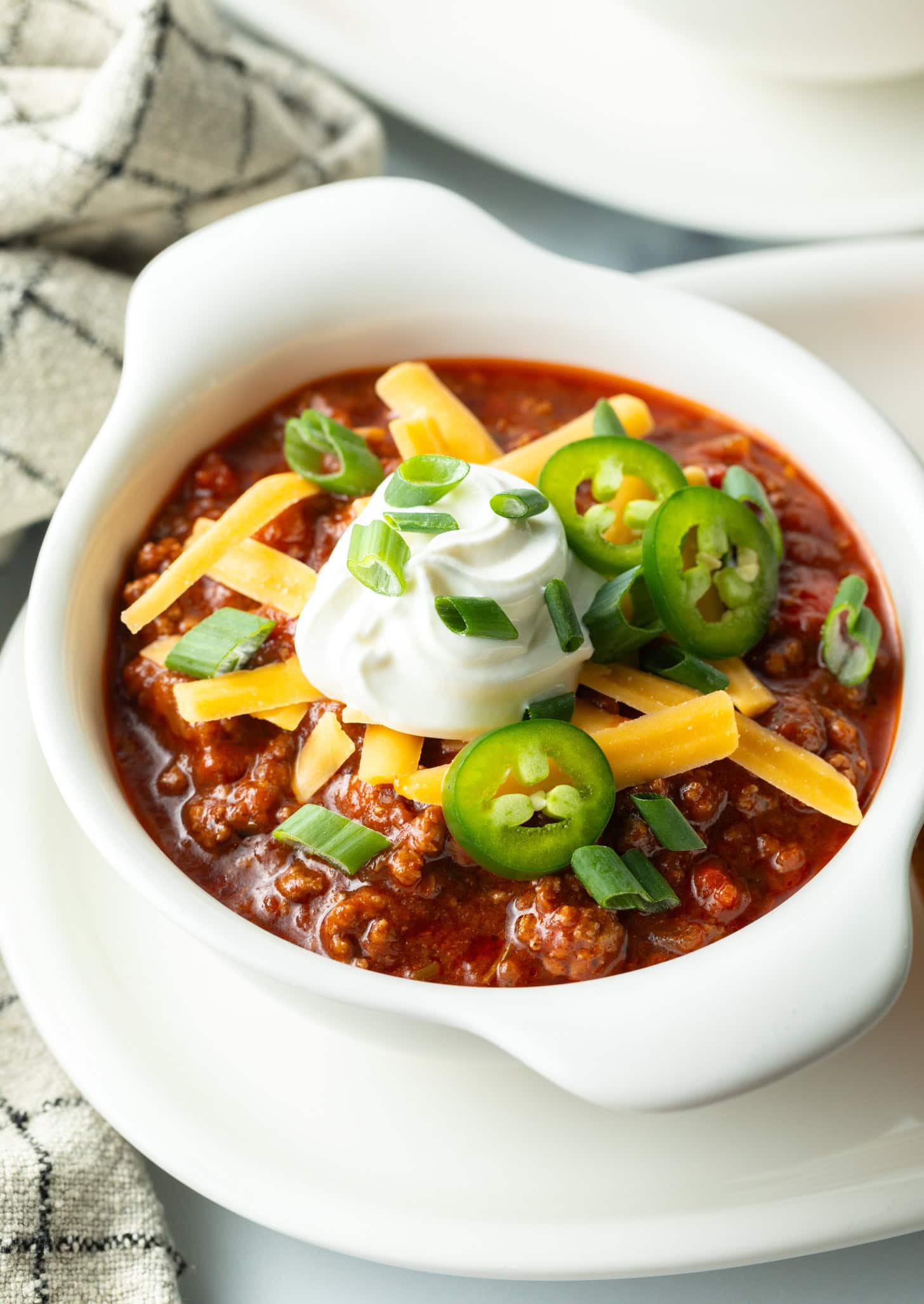 Top down view of a white bowl of beanless chili topped with shredded cheese, jalapeno slices, sour cream and chopped green onions.