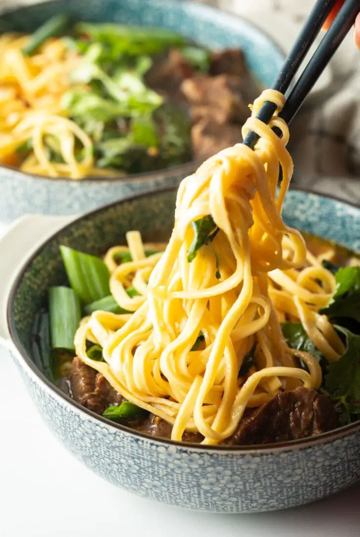 Hand mit Stäbchen greift Nudeln aus der Schüssel mit Rindfleischsuppe.