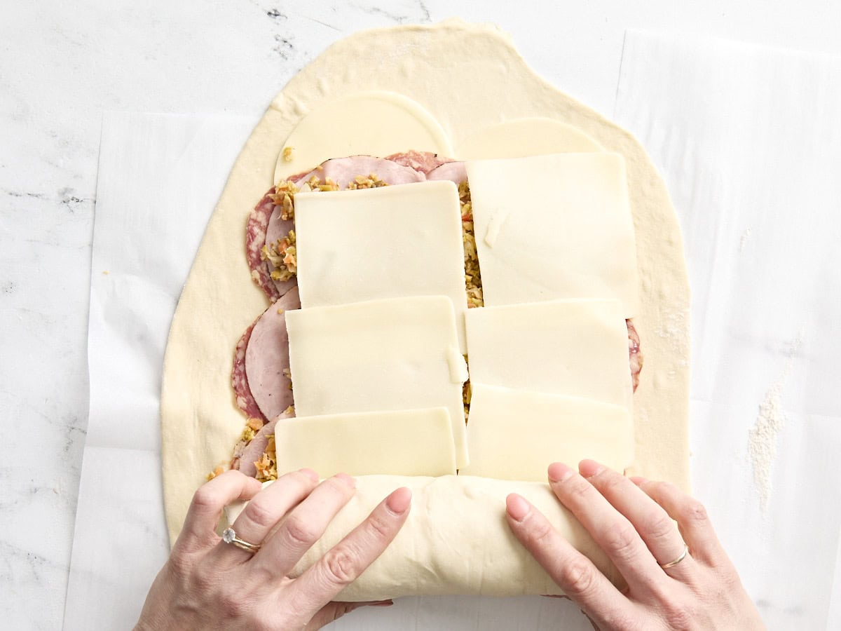 Hände rollen den Pizzateig über die Füllungen für Muffaletta-Roll-ups