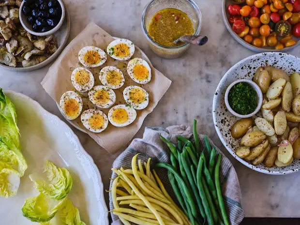 Zutaten für eine Nicoise auf einer Theke angeordnet, darunter hartgekochte Eier, grüne Bohnen, Kartoffeln, Oliven, Salat