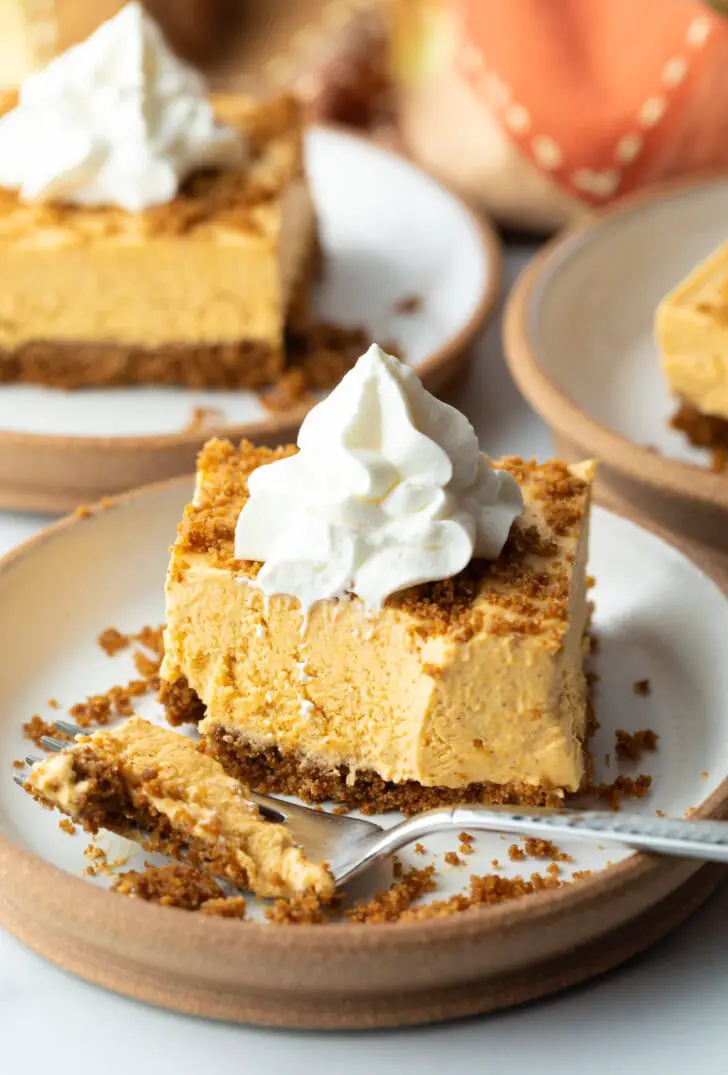 Quadratischer Kürbiskuchen-Käsekuchen auf einem weißen Teller, garniert mit Schlagsahne. Auf dem Teller liegt auch eine Gabel mit einer Kugel des Desserts.