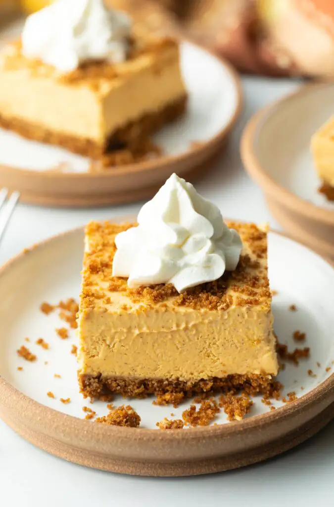 Square of pumpkin pie cheesecake on a white plate, topped with whipped cream.