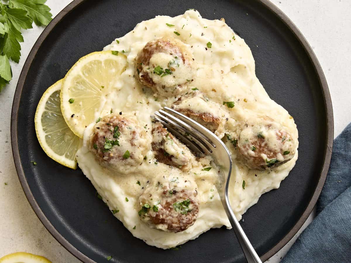 Draufsicht auf Hähnchenfleischbällchen mit Sahnesauce, serviert auf einem Teller mit Kartoffelpüree, und einer Gabel, die ein Fleischbällchen in zwei Hälften schneidet.